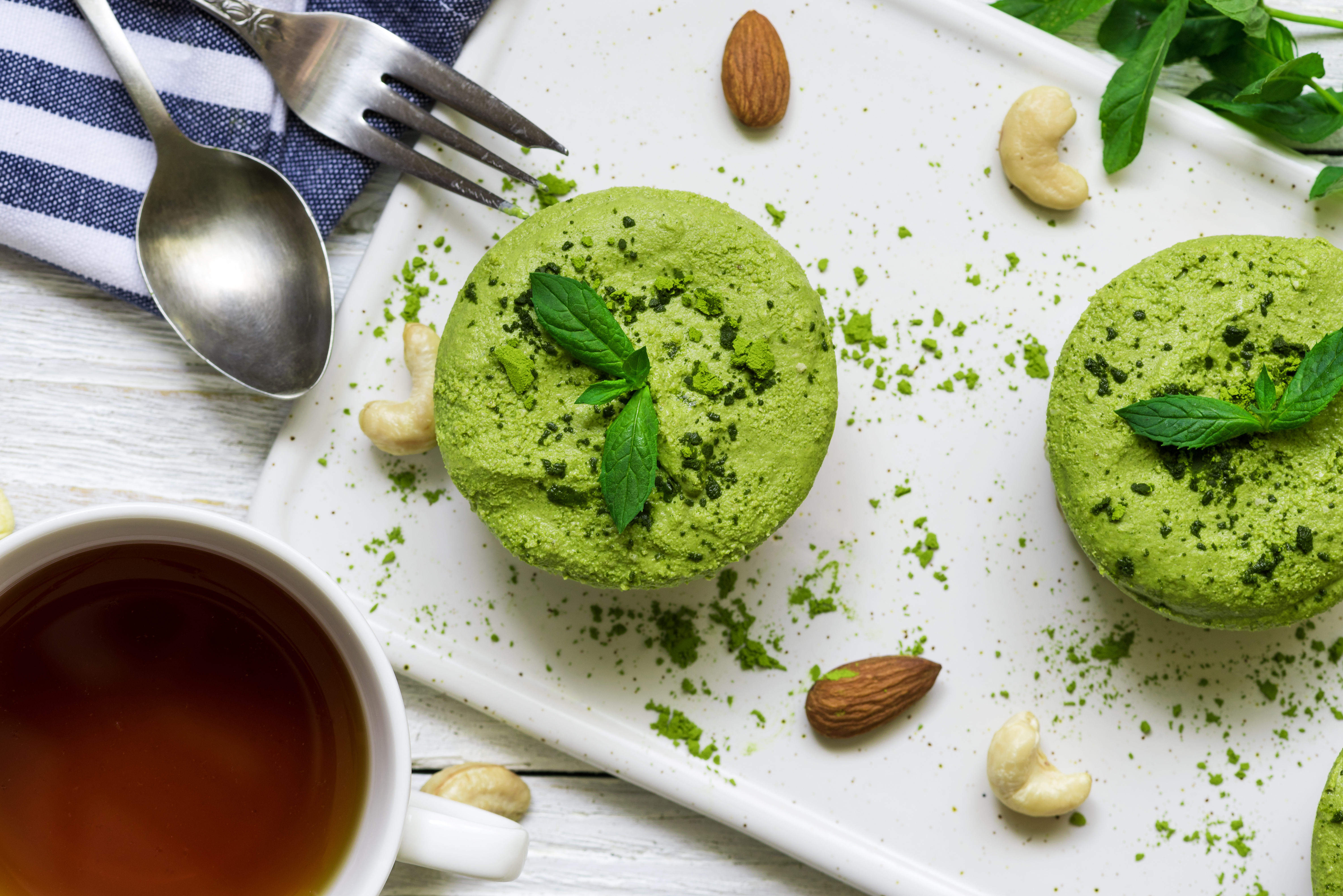 green matcha and banana vegan raw cheesecakes with mint and nuts over white wooden table with cup of tea and spoon. healthy delicious breakfast. top view